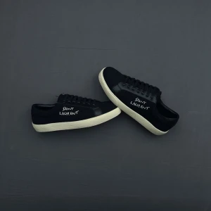 Svarta Saint Laurent sneakers - Svarta sneakers från Saint Laurent med vit textlogga på sidan. Skorna har en låg silhuett, svart ovandel i canvas och läderdetaljer samt vit gummisula. Klassisk snörning framtill och stilren design som passar till det mesta.