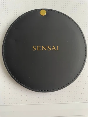 Rund sminkspegel från Sensai - Stilren rund sminkspegel från Sensai med svart utsida och guldfärgad knappdetalj. Insidan är röd och spegeln är perfekt att ha i väskan. Elegant design med Sensai-logga i guld på framsidan. Praktiskt och snyggt tillbehör för sminkrutinen.