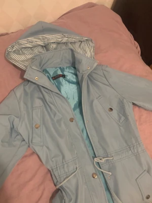 Ljusblå regnjacka med randig huva - Snygg ljusblå regnjacka  med dragsko i midjan och stora fickor med tryckknappar. Jackan har en huva med vitt och blått randigt foder och stängs med både dragkedja och knappar. Perfekt för regniga dagar.