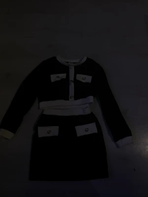 Barn klänning sett  - Snyggt tvådelat set i svart och vitt med långärmad crop top och matchande kjol. Setet har dekorativa fickor och guldiga knappar framtill. Materialet ser ut att vara mjukt och stretchigt, perfekt för en trendig look.