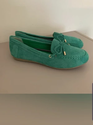 Gröna mockaloafers med rosett - Snygga gröna loafers i mocka med dekorativ rosett framtill och guldfärgade metalldetaljer. Skorna har en klassisk, låg sula i gummi och stilren design som passar till många olika outfits. Ej äkta mocka skinn