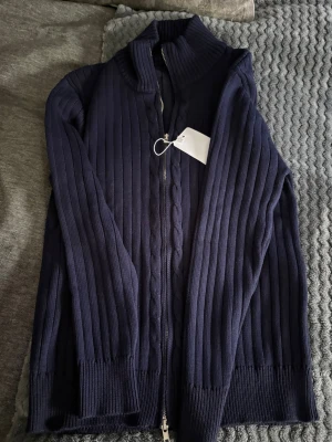 Mörkblå ziptröja maison margiella  - Mörkblå stickad ziptröja med hög krage och hel dragkedja framtill. Tröjan har en smal passform och ribbade detaljer längs ärmar och kropp. Perfekt för lager-på-lager och enkel att matcha med olika outfits.