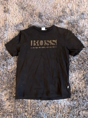 Svart BOSS t-shirt med gulddetaljer - Snygg svart t-shirt från BOSS med guldfärgat logotryck på bröstet. Klassisk rund halsringning och korta ärmar. Tillverkad i mjuk bomull som känns skön mot huden. Perfekt för dig som vill ha en stilren och trendig look.