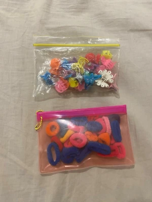 Färgglada hårsnoddar och hårklämmor - Säljer ett mixat set med elastiska hårsnoddar och små hårklämmor i olika färger som rosa, blå, gul, orange och lila. Perfekt för att fixa roliga frisyrer eller sätta färg på håret. Allt kommer i två praktiska plastväskor för enkel förvaring.