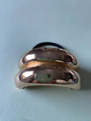 Guldig hårklämma med elastiskt band - Snygg håraccessoar i form av en guldig hårklämma med rundad, metallisk yta och ett svart elastiskt band. Perfekt för att skapa en trendig look och hålla håret på plats. Klämman har en modern, chunky design som sticker ut.