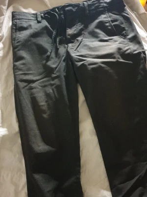 Grå kostymbyxor med normal passform - Säljer ett par grå kostymbyxor med lite street, resårbandband vid ankeln, normal passform. Byxorna har snedställda fickor fram och bälteshällor i midjan. Perfekta till skjorta eller stickad tröja för en clean look.