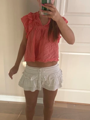 Korall volangblus från neo noir  - Säljer en söt rosa topp från neo noir med volangdetaljer och smock vid axlar och bröst. Blusen är ärmlös och har en luftig, croppad passform som är perfekt för varma dagar. Materialet är lätt och skrynkligt vilket ger en avslappnad look.