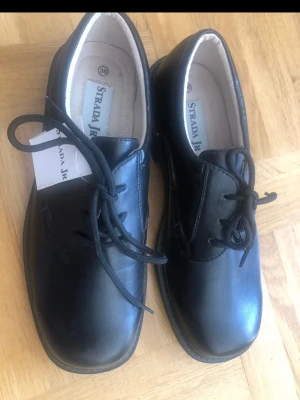 Svarta loafers från Strada JR 36 nya - Klassiska svarta loafers från Strada JR i storlek 36. Skorna har slät ovandel i läderimitation, rund tå och snörning framtill. Snygg och enkel design som passar till många olika stilar. Bekväm innersula och diskret sula i svart.