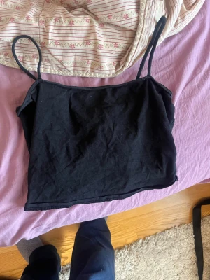 Svart linnetopp med smala axelband - Enkel och stilren svart linnetopp med smala axelband. Perfekt att matcha med jeans eller kjol för en clean look. Tillverkad i mjuk bomull som känns skön mot huden. Croppad modell som ger en trendig vibe.