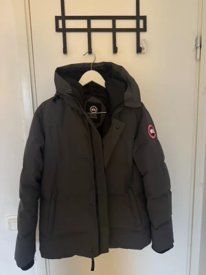 Mörkgrå dunjacka från Canada Goose - Snygg mörkgrå dunjacka från Canada Goose med huva och klassisk logotyp på ärmen. Jackan har dragkedja och knappar framtill, två stora fickor och en varm, puffig design. Perfekt för kalla vinterdagar och riktigt skön att bära.  LÄGG ETT PRISFÖRSLAG