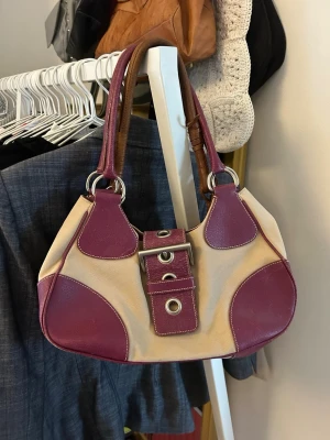 Vintage Prada handväska - Snygg handväska i beige tyg med vinröda detaljer och handtag i skinnimitation. Väskan har två stora ringar i metall, dekorativa spännen framtill och kontrastsömmar. Perfekt accessoar för att lyfta din outfit.
