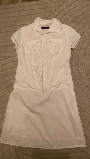 Vit skjortklänning med kort ärm - Snygg vit skjortklänning med korta ärmar och krage. Klänningen har knappar framtill, två bröstfickor och markerad midja för en feminin siluett. Perfekt till sommaren och enkel att styla med accessoarer. StockholmStyle