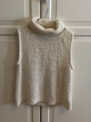 Vit stickad väst från InWear - Mysig vit stickad väst från InWear med hög polokrage. Västen är ärmlös och har en loose passform, perfekt att styla över en skjorta eller långärmad tröja. Materialet är mjukt och fluffigt, vilket ger en chill och trendig vibe.