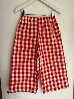 Rödrutiga byxor med resårmidja - Unika rödrutiga handsydda byxor med bred passform och resår i midjan. Tillverkade i ett mjukt bomullstyg med klassiskt rutmönster i rött och vitt. Perfekta för en avslappnad och trendig stil. Märkta med 'Handmade' på sidan. Passar 18-24, 92. 