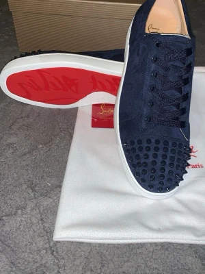 Marinblå Christian Louboutin sneakers - Snygga marinblå sneakers från Christian Louboutin med klassiska röda sulor och svarta nitar på tån. Skorna är låga och har ovandel i mocka, vita yttersulor och svarta skosnören. Perfekta för dig som vill sticka ut med exklusiv design.