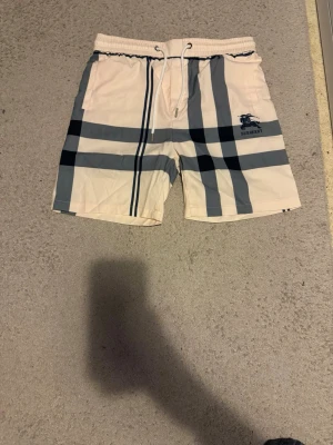Beige-rutiga badbyxor från Burberry - Säljer ett par snygga badbyxor från Burberry i beige med klassiskt rutmönster i svart och grått. De har resår och dragsko i midjan samt Burberry-logga på vänster ben. Perfekta för stranden eller poolen, riktigt stilrena och trendiga.
