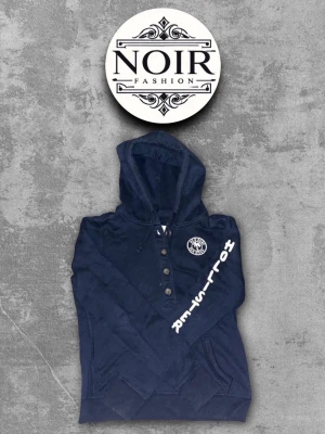 Mörkblå hoodie från Hollister  - Snygg mörkblå hoodie med huva från Hollister. Hoodien har knappar framtill, vit tryckt logga på bröstet och text på ena ärmen. Perfekt för dig som gillar streetstyle och vill ha något bekvämt och trendigt. Materialet känns mjukt och skönt.