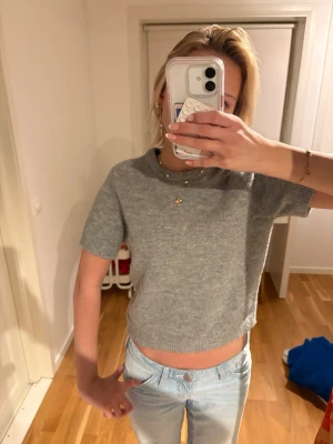 Grå stickad topp - Grå stickad tröja med korta ärmar och rund hals. Enkel och stilren design som passar perfekt till jeans eller kjol. Mjuk och bekväm, perfekt för dig som gillar en avslappnad men trendig look.