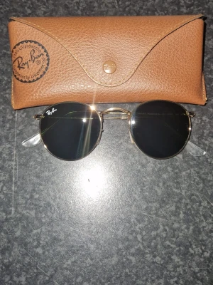 Runda solglasögon från Ray-Ban - Snygga runda solglasögon från Ray-Ban med guldfärgad metallram och mörka linser. Medföljer ett brunt fodral i skinn. Perfekt för dig som vill ha en klassisk och tidlös look.