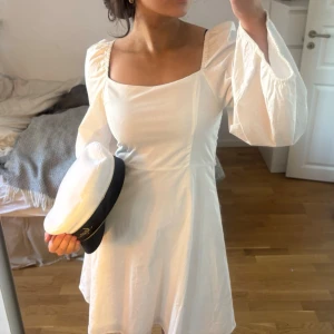 Vit kort klänning med vida ärmar - Säljer en vit kort klänning med fyrkantig ringning och vida, långa ärmar. Klänningen har en lätt utsvängd passform och är perfekt för student eller sommar. Enkel och stilren design utan synliga detaljer eller mönster. Sprettatupp resonen i armarna för en mordernare look 