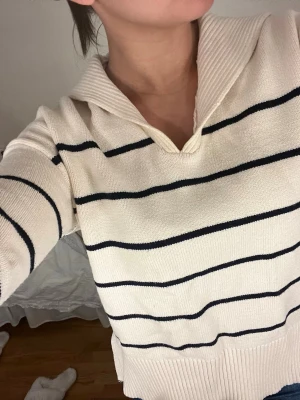 Randig stickad tröja med krage - Vit beige stickad tröja med svarta horisontella ränder och bred krage. Tröjan har v-ringning, långa ärmar och ribbade muddar samt nederkant. Perfekt för en avslappnad och trendig look.