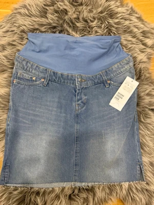 Blå gravidjeanskjol med stretch - Säljer en blå jeanskjol med mjuk stretchig mudd upptill, perfekt för gravidmage. Kjolen har klassiska jeansdetaljer som fickor och bälteshällor samt rå kant nertill. Enkel och snygg att matcha till olika outfits.