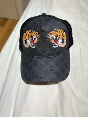 Svart Gucci keps med tigertryck - Snygg svart keps från Gucci med två broderade tigrar framtill och diskret GG-mönster över hela kepsen. Baksidan har meshmaterial för extra ventilation och justerbar rem. Perfekt för dig som vill sticka ut med en exklusiv accessoar.