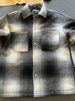 Svartvit rutig overshirt Jack & Jones - Overshirt från Jack & jones i svartvitt rutigt mönster med inslag av beige. Två stora bröstfickor med lock, klassisk krage och tryckknappar framtill. Tjockt och mjukt material som ger en avslappnad look. Perfekt lager på lager-plagg för kyligare dagar.