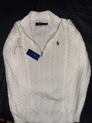 Vit kabelstickad tröja Polo Ralph Lauren - Kabelstickad vit tröja från Polo Ralph Lauren med halv dragkedja och broderad logga på bröstet. Tröjan har ribbade muddar och hög krage, vilket ger en klassisk och stilren look. Perfekt för dig som gillar tidlös design och premiumkänsla.
