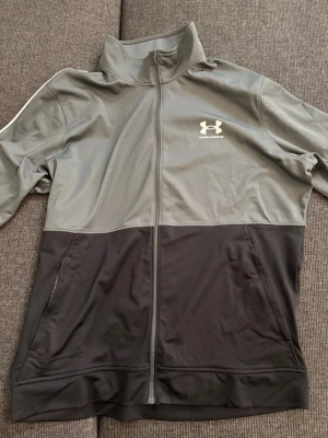 Under Armour - Snygg träningsjacka från Under Armour i grått och svart med hel dragkedja och hög krage. Jackan har vit logga på bröstet och en sportig design med tvådelad färg. Tillverkad i ett mjukt och stretchigt funktionsmaterial. Passar som L och skriv i dm för fler frågor.