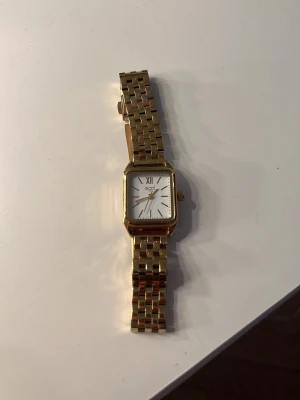 Guldfärgad klocka Regal - Stilren klocka från Regal med fyrkantig urtavla och guldfärgad länk i metall. Urtavlan är vit med romerska siffror och tunna visare. En tofs har stött emot klockan så lite av färgen har försvunnit men inget man ser