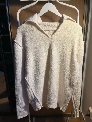 Vit stickad hoodie med v-ringning - Säljer en krämvit stickad hoodie med v-ringning och luva. Tröjan har en avslappnad passform, långa ärmar och ribbade muddar. Materialet är mjukt och strukturerat, perfekt för lager på lager. Passar dig som gillar en clean och enkel stil.