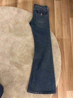 Blå bootcut jeans med fickdetalj - Snygga blå bootcut jeans med klassisk femficksdesign och en extra ficka med knapp bak. Jeansen har en lätt tvättad look framtill och är tillverkade i ett mjukt denimtyg. Perfekta för dig som gillar en avslappnad men trendig stil.