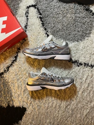 P6000 Nike - Snygga sneakers från Nike i silver och vitt med svarta detaljer. Skorna har en sportig design med mesh och syntetmaterial, samt en vit och svart sula. Perfekta för dig som gillar en futuristisk och clean stil.