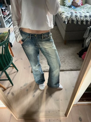 Blå bootcut jeans från Replay - Säljer ett par ljusblå bootcut jeans från med slitningar och kontrastsömmar. Jeansen har låg midja, fyra fickor och broderade detaljer på bakfickorna. Perfekta för en avslappnad och trendig look.