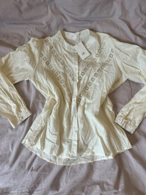 Vit blus med spetsdetaljer - Söt vit blus med knappar framtill och dekorativa spetsdetaljer i V-form över bröstet. Blusen har rund halsringning och långa ärmar med lätt puff vid axlarna. Tillverkad i ett tunt, luftigt material som känns fräscht och somrigt.