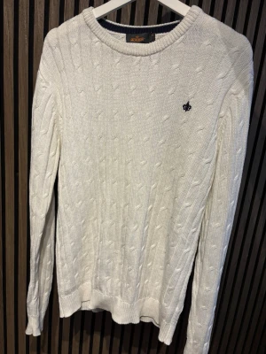 Vit kabelstickad tröja Morris - Kabelstickad vit tröja från Morris med rund halsringning och diskret broderad logga på bröstet. Tröjan har långa ärmar och ribbade muddar vid ärmslut och nederkant. Perfekt för en clean och klassisk stil.