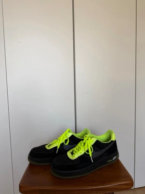 Nike Air Force 1 - Säljer ett par Nike Air Force 1 sneakers i svart mocka med neongula detaljer, snören och foder. Klassisk låg modell med tydlig AIR-text på sulan och ikonisk swoosh på sidan. Perfekta för dig som vill sticka ut med färg och stil. 