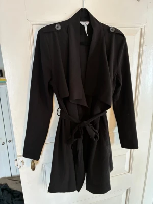 Svart trenchcoat med knytskärp OBJECT - Stilren svart trenchcoat med långa ärmar, bred krage och knytskärp i midjan. Jackan har axelklaffar med dekorativa knappar och ett mjukt, följsamt tyg som ger en avslappnad look. Perfekt för dig som vill ha en klassisk men modern trench. Passar S snarare än M