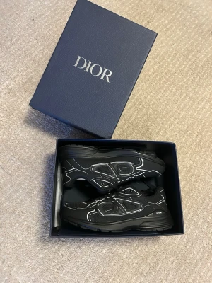 Svarta Dior B30 sneakers - Snygga svarta Dior B30 sneakers med vita detaljer och reflekterande linjer. Skorna har en sportig siluett, mesh och läderpaneler samt tydlig Dior-logga på sidan och plösen. Perfekta för dig som vill ha en exklusiv och trendig look.