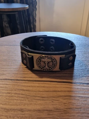 Armband vintage Tors hammare - Svart läderarmband med ett detaljerat Tors hammar i metall. Armbandet har justerbara tryckknappar för att passa olika handledsstorlekar. Storlek 23 cm långt. Bredd 3 cm.