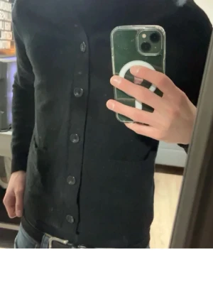 Svart kofta med knappar - Stilren svart kofta med stora knappar framtill och två fickor. Klassisk rak passform och långa ärmar, perfekt att slänga över en t-shirt eller skjorta. Passar till jeans eller chinos för en clean look.