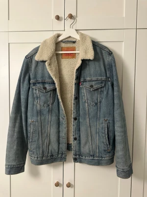 Levi's blå jeansjacka med foder - Klassisk blå jeansjacka från Levi's med mysigt teddyfoder och krage i offwhite. Jackan har knappar framtill, två bröstfickor med lock och två sidofickor. Perfekt oversized look och passar till lager-på-lager-stil.