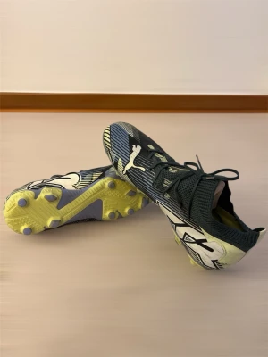 Puma fotbollsskor svart/gul - Säljer ett par snygga Puma fotbollsskor i svart och gul med vita detaljer och coolt grafiskt mönster. Skorna har snörning, stickad strumpliknande krage och platt sula med dobbar för bra grepp på planen. Perfekta för dig som vill sticka ut på matchen!
