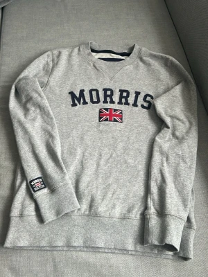 Grå sweatshirt från Morris - Snygg grå sweatshirt från Morris med mörkblå text och brittisk flagga på bröstet. Tröjan har rund halsringning, ribbade muddar och en patch på ärmen. Tillverkad i mjukt bomullsmaterial som är perfekt för en avslappnad stil. 