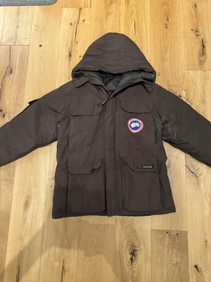 Brun dunjacka från Canada Goose - Säljer en brun dunjacka från Canada Goose med huva och klassisk logga på bröstet. Jackan har stora fickor framtill, dragkedja och är perfekt för kalla vinterdagar. Fodrad med dun för extra värme och har en stilren, funktionell design.