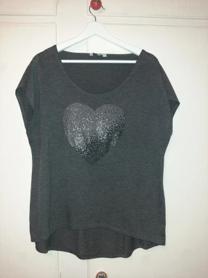Mörkgrå topp med glittrigt hjärta från New Look - Snygg mörkgrå topp med ett stort glittrigt hjärta framtill. Toppen har rund halsringning och korta ärmar, perfekt för en chill och trendig look. Materialet känns mjukt och bekvämt mot huden.