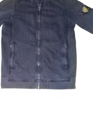 Mörkblå ziptröja från Stone Island - Mörkblå ziptröja från Stone Island med ikonisk patch på ärmen. Tröjan har två fickor framtill, ribbade muddar och dragkedja hela vägen. Tillverkad i mjuk bomull för en avslappnad och snygg look. PRIS KAN DISKUTERAS!!! 