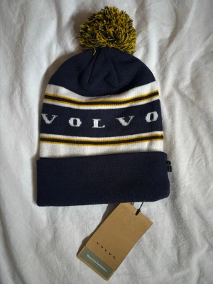 Volvo Marinblå Mössa - Snygg marinblå mössa från Volvo med gul och vit rand, samt stor tofs på toppen. Påminner om Åre/Mintu mössorna 💕 säljes pga ingen användning 