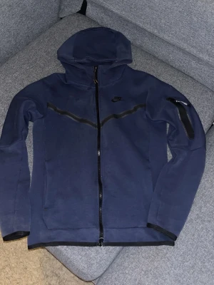 Mörkblå Nike Tech Fleece hoodie - Nike Tech Fleece hoodie i mörkblått med hel dragkedja och svarta detaljer. Huva, fickor på sidorna och en extra ficka med dragkedja på ärmen. Tillverkad i mjukt och bekvämt fleecematerial, perfekt för en sportig och avslappnad stil.